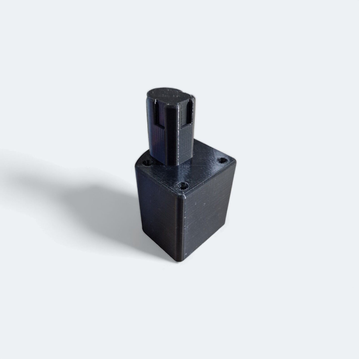 A compact black plastic cube with a cylindrical top, designed as an adaptable holder perfect for Ryobi One+ tools, is showcased on a light gray background. This efficient storage solution casts a soft shadow to the left, emphasizing its usefulness in keeping tools organized. The product is identified as the Deep Mount by the brand Battery.