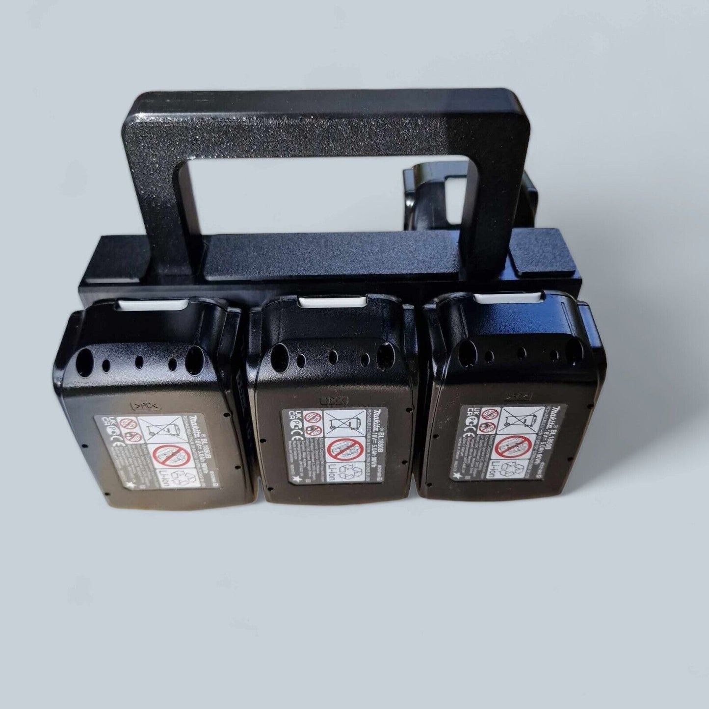 A sleek black Battery brand Basket Carrier Caddy Holder for Makita, featuring three storage compartments each fitted with a black battery, is mounted on a plain light gray background. The batteries display white labels accented with diagrams and caution symbols, resembling a modern battery basket.